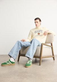 Pulover crem cu logo-ul "GANT", blugi albaștri deschiși, adidași verzi cu accente albe, așezat pe un scaun bej cu cadru din lemn.