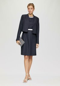 Robe bleu marine foncé avec une silhouette ajustée, accents blancs et une veste courte. Le modèle tient une pochette grise texturée, portant des sandales blanches à brides.