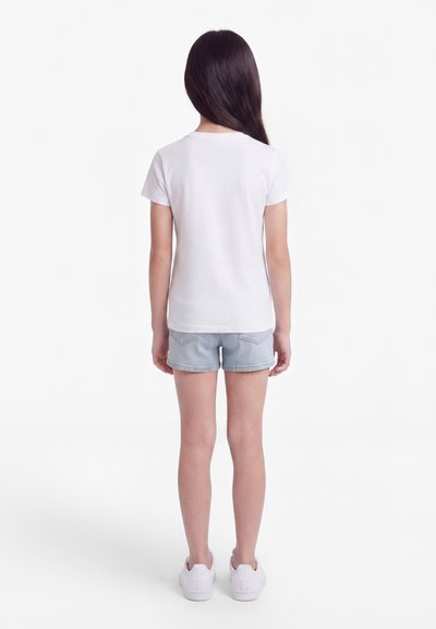 Dieťa s dlhými tmavými vlasmi, ktoré má na sebe obyčajné biele tričko, svetlomodré rifľové kraťasy a biele tenisky, stojace chrbtom.