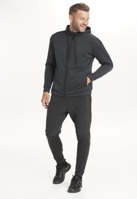 Musta vetoketjullinen huppari, jossa on teksturoitu kangas, säädettävä nyörillinen huppu, yhdistettynä vartalonmyötäisiin mustiin housuihin ja mustiin urheilujalkineisiin.