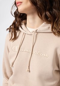 Felpa beige realizzata in morbido tessuto con un logo "Champion" ricamato. Presenta un cappuccio regolabile con lacci e una finitura testurizzata.