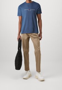 Blue cotton t-shirt with "TOMMY HILFIGER" logo, beige tailored trousers, and white sneakers. Holding a black quilted shoulder bag.