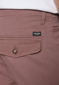 Detailní záběr na zadní kapsu fialových kalhot s klopou a knoflíkem, obsahující černou nálepku "Jack & Jones EST. 1990" a viditelné švy.