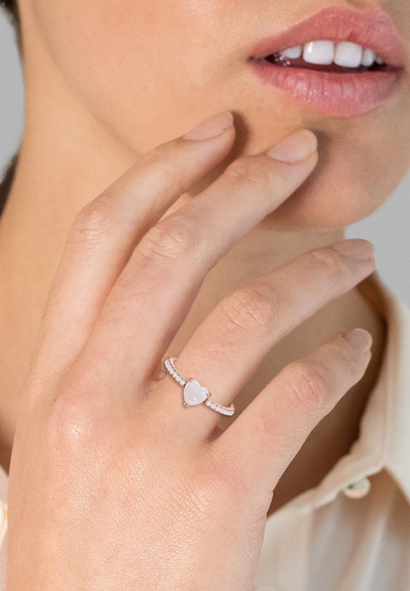 Zlato-rozno prstan s središnjim kamnom v obliki srca iz opala, okrašenim z majhnimi prozornimi kristali ob obroču. Gladek, poliran zaključek.