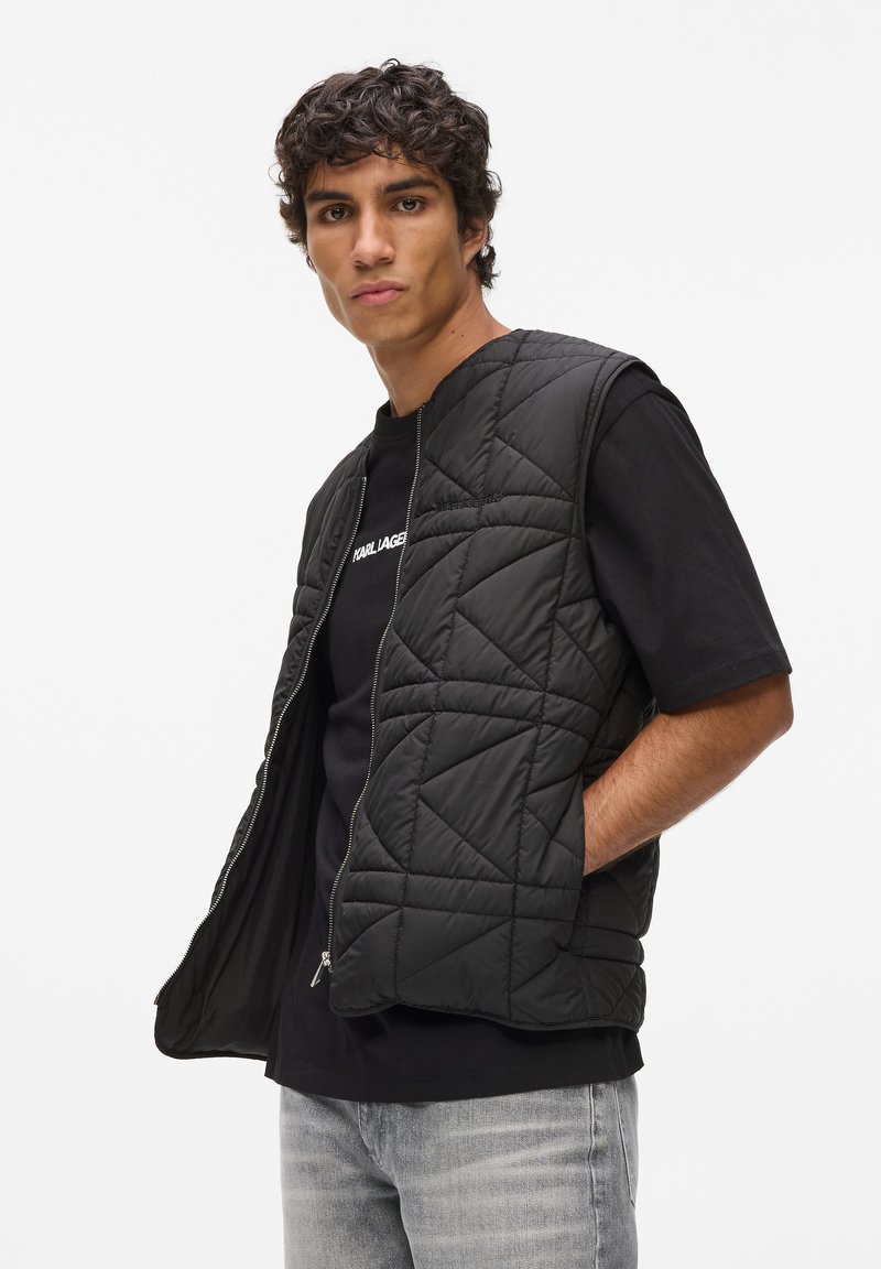 Zwarte doorgestikte bodywarmer met ritssluiting, voorzien van een hoge halslijn en open armsgaten, gedragen over een zwart T-shirt en lichtgrijze jeans.