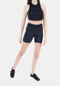Top corto nero abbinato a pantaloni corti aderenti neri. Entrambi i capi hanno una texture liscia. Indossati con scarpe da atletica nere con suole bianche.