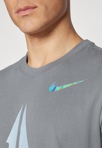 Camiseta de algodón gris con cuello redondo, un logo del swoosh de Nike reflectante y multicolor, y un diseño gráfico simplificado en la parte frontal.