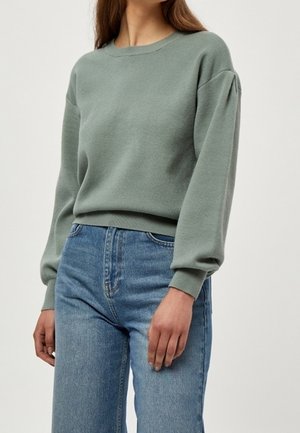 Žena v světle zeleném svetru s dlouhými rukávy a nafouklými rameny a modrých džínech s vysokým pasem na jednobarevném pozadí.