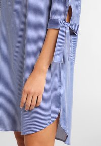 Gestreiftes Hemdkleid in Blau und Weiß, mit hochgekrempelten Ärmeln mit Bändern, Seitenschlitzen und einer lockeren, entspannten Passform. Aus Baumwollmaterial.