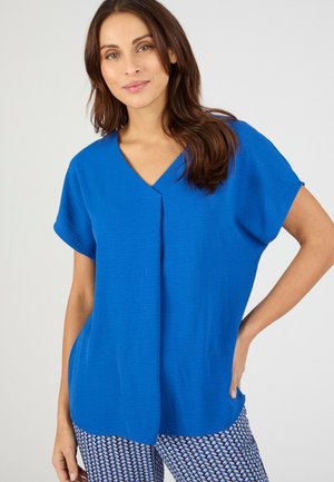 Mujer con una blusa azul de manga corta y cuello en V holgada, y pantalones estampados en azul y blanco, posando con una mano en la cadera.