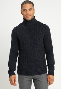 Suéter de cuello alto azul marino con un patrón de punto trenzado texturizado, que presenta puños y dobladillo acanalados. Combinado con pantalones grises, vista recortada.