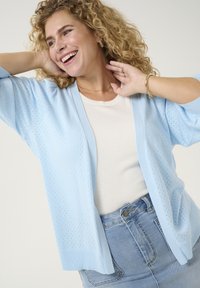 Light blue knitted cardigan with a loose fit, featuring a perforated texture, open front, and ribbed cuffs, paired with a white shirt and denim skirt.
