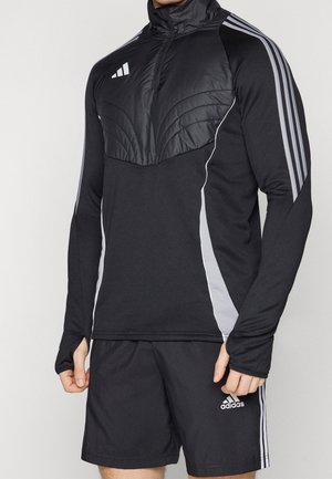 Homme portant une veste de sport noire Adidas avec un panneau matelassé sur la poitrine et un short assorti avec des bandes blanches sur les manches et les côtés.