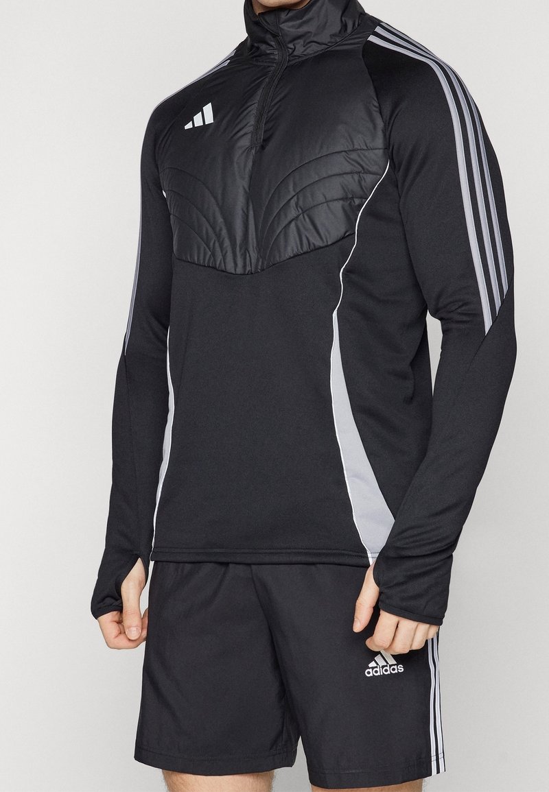 Homme portant une veste de sport noire Adidas avec un panneau matelassé sur la poitrine et un short assorti avec des bandes blanches sur les manches et les côtés.