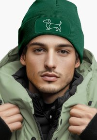 Green knitted beanie featuring a white dachshund graphic, paired with a light green puffer coat, showcasing a layered look and texture.
