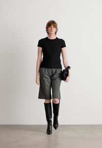 Svart strukturert topp med kort erm, grå høytlivsshorts og knelang svarte støvler. Modellen holder en svart håndveske i venstre hånd.