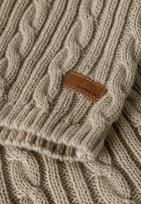 Beżowy sweter dzianinowy z wzorem w warkocze i brązową skórzaną metką. Posiada ściągacze na rękawach i miękkie, teksturowane wykończenie.