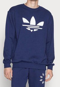 Granatowa bluza z kapturem z białym logo Adidas, przedstawiającym trzy liście i paski. Wykonana z miękkiego, fakturowanego materiału z prążkowanymi mankietami.