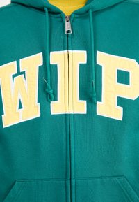 Haut à zip teal avec lettering jaune "WIP". Comprend une grande poche avant et des cordons de serrage. Fabriqué en tissu doux avec une texture lisse.
