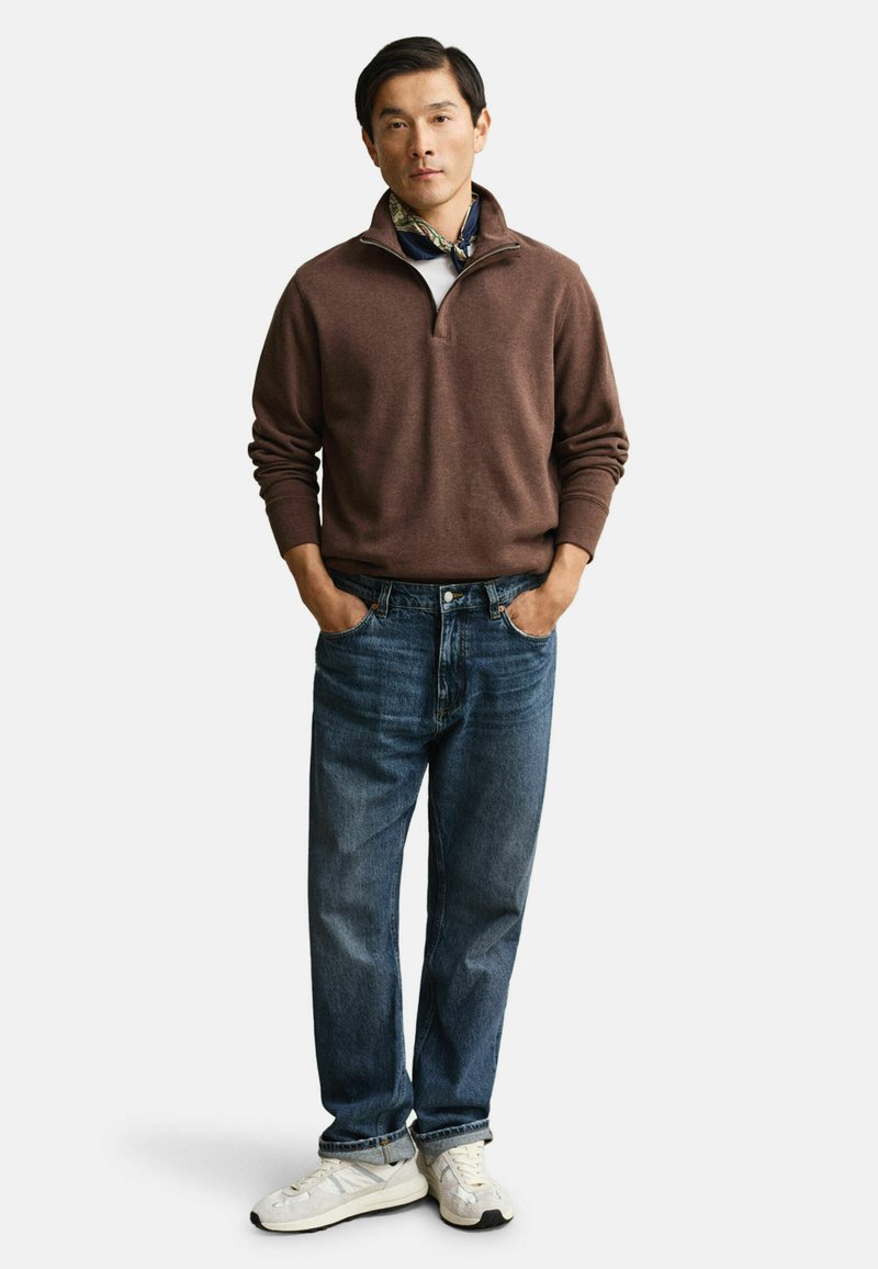 Brun zip-up tröja, vit skjorta under, blå jeans av denim och vita sportskor. Modellen står med händerna i fickorna.