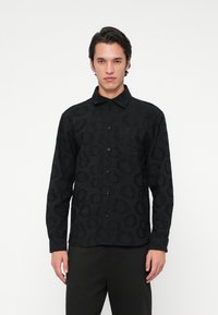 Egy férfi fekete gombos ingben, amely finom körkörös mintákkal díszített, és fekete nadrágban áll egy sima fehér háttér előtt.