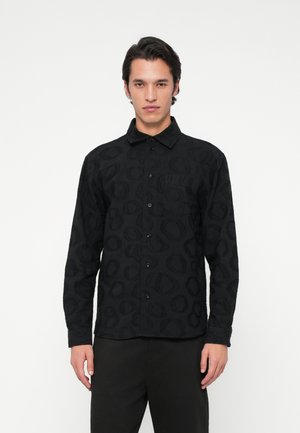 Mees mustas nööpidega särgis, millel on peened ringikujulised mustrid, ja mustades pükstes, seistes lihtsa valge tausta ees.