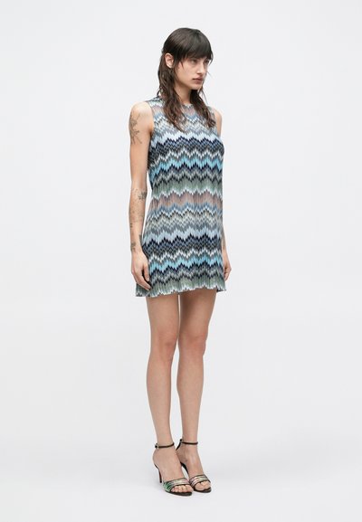 Žena s dlhými tmavými vlasmi a tetovaniami na rukách má na sebe priesvitné minišaty s viacfarebným vzorom cik-cak a sandále na vysokých podpätkoch s remienkom okolo členka.