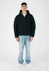 Giacca a vento nera con cappuccio, con chiusura lampo frontale e tasca sul petto. Indossata con jeans in denim blu chiaro e sneakers bianche.