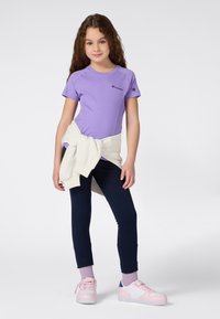 Jeune fille aux longs cheveux bouclés portant un t-shirt violet Champion, un legging bleu marine, des chaussettes roses, des baskets et un sweat-shirt blanc noué autour de la taille.