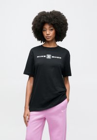 Μαύρο t-shirt Nike Shox με κοντά μανίκια, με λευκό λογότυπο στο εμπρός μέρος. Φορεμένο με ανοιχτό ροζ φόρμας, το ύφασμα φαίνεται μαλακό.