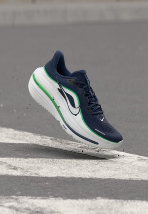 Chaussure de course bleue et blanche avec une bordure verte flottant au-dessus de l'asphalte avec des marquages blancs, petites particules de saleté visibles autour de la semelle.