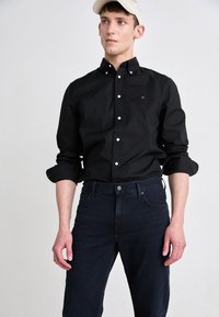 Black button-up shirt featuring a small logo, with rolled sleeves, paired with dark jeans. Model wears a light-colored cap.