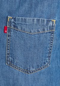 Nära bild av denimtyg med en blå bakficka som har gul tråd i sömmen och en liten röd etikett nära fickans kant.