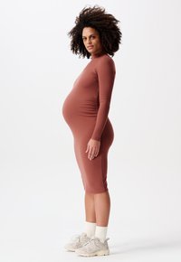 Umstands Kleid aus geripptem Terrakotta-Stoff, mit taillierter Passform, langen Ärmeln und knielangem Saum. Getragen mit hellen Sneakers.