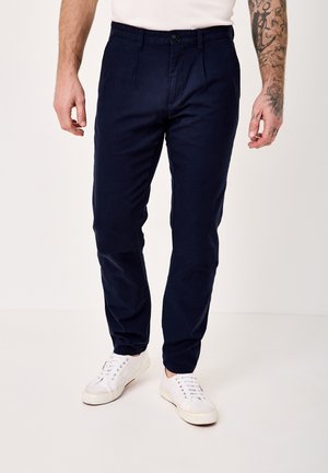 Man wearing navy blue trousers and white sneakers, standing with tattooed right arm visible, against a plain light background.