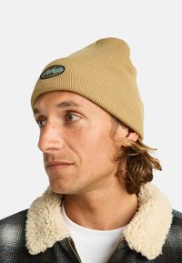 Beanie a coste beige con patch logo, indossato con una giacca con collo in felpa. Dettaglio del profilo della testa. Tessuto texture visibile.
