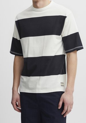 Homme portant un t-shirt à manches courtes avec de larges rayures horizontales noires et blanches, assorti à un pantalon foncé, debout devant un fond uni.