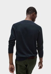 Námořnicky modrý pletený svetr s kulatým výstřihem, raglánovými rukávy a žebrovanými manžetami. Měkká textura s minimálním zdobením, nošený přes olivově zelené kalhoty.