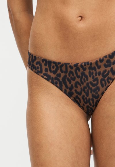 Femme portant un bas de bikini marron et noir imprimé léopard montrant le ventre et le haut des cuisses sur fond blanc.