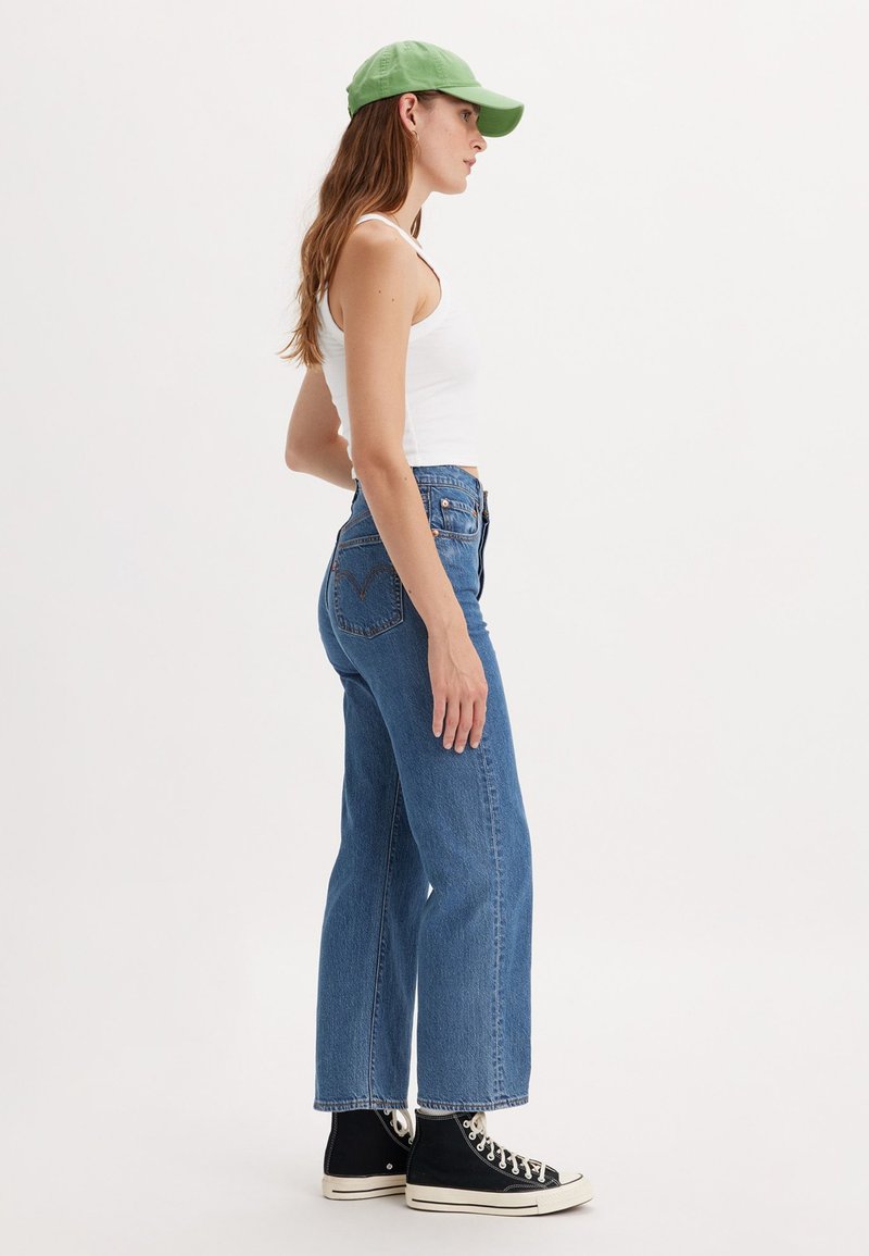 Jean en denim bleu taille haute avec une coupe droite, délavé clair, coutures visibles et un logo subtil sur la poche arrière. Associé à un débardeur blanc et une casquette verte.
