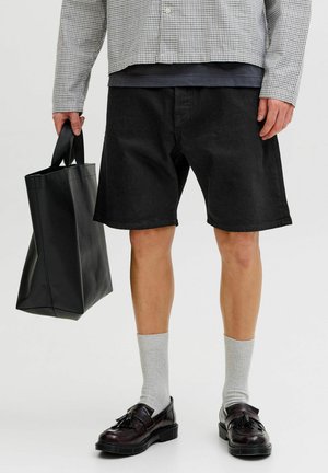 Henkilö, jolla on mustat shortsit, ruudullinen paita, harmaat sukat, mustat loafers-kengät, vasemmassa kädessä suuri musta olkalaukku.