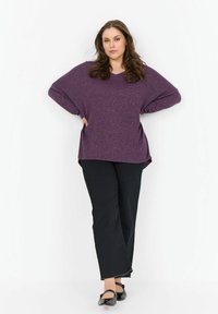 Lila, oversize V-nyakú pulóver, texturált felülettel, fekete nadrággal és fekete lapos cipőkkel párosítva, oldalsó zsebekkel.