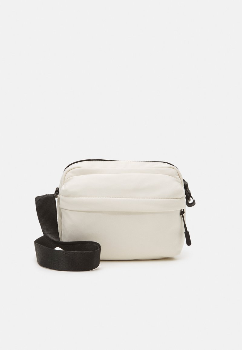 Hvid stof crossbody taske med sort justerbar rem, der har en lynlåset hovedrum og en frontlomme til opbevaring.