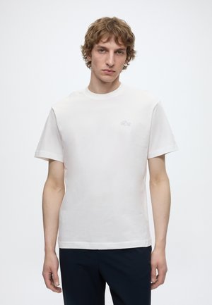 Jeune homme aux cheveux bouclés portant un t-shirt blanc uni à manches courtes et un pantalon sombre sur fond blanc