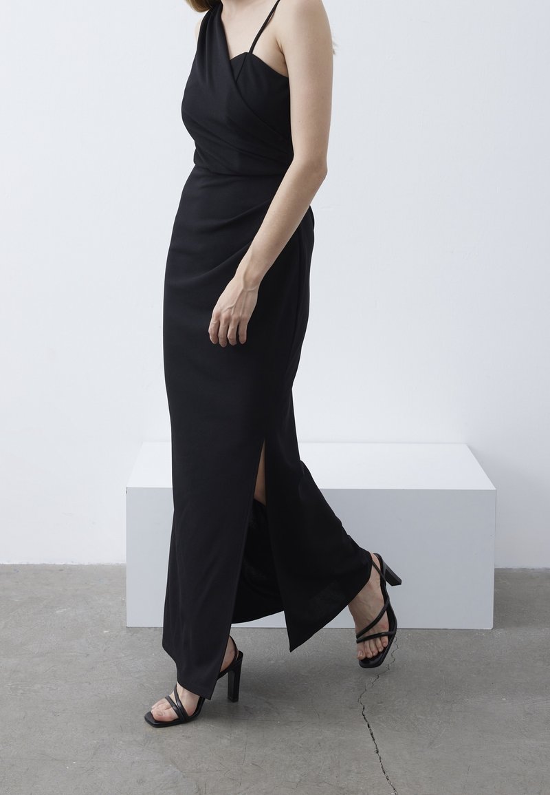 Robe noire, longue, avec un design enroulé, de petites fentes sur les côtés et des fines bretelles. Portée avec des sandales à talons hauts noires avec lanières.