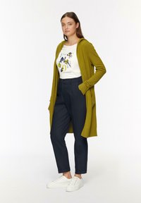 Long cardigan vert olive avec une capuche, porté par-dessus un t-shirt graphique blanc. Associé à un pantalon bleu marine et des baskets blanches. Look décontracté.