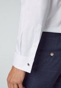 Witte overhemd met een gestructureerde afwerking, voorzien van lange mouwen en een ronde manchet, accentueert door een zilveren manchetknopen. Marineblauwe geruite broek zichtbaar.