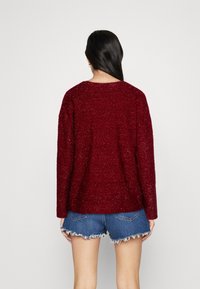 Pull en maille bordeaux à coupe ample et manches longues, associé à un short en jean effiloché. La texture semble douce et épaisse.