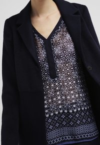 Czarna, teksturowana marynarka noszona na przezroczystej, granatowej bluzce z geometrycznym wzorem i dekoltem w kształcie litery V. Posiada detal w postaci guzików oraz warstwowy krój.