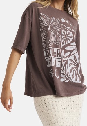 Femme portant un t-shirt marron ample avec un motif floral et lunaire blanc et le mot "Billabong,", associé à une jupe texturée crème.